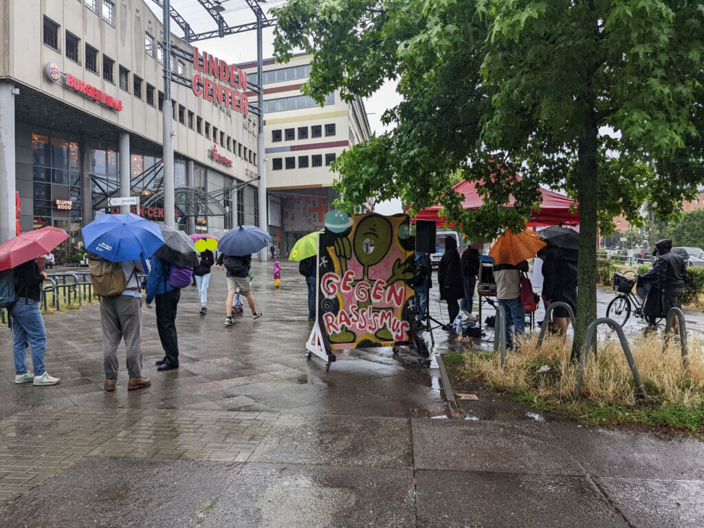 The square in front of the shopping center "Lindencenter" on a rainy day. Here is a pavilion, a photo wall against racism and some people with colorful umbrellas.