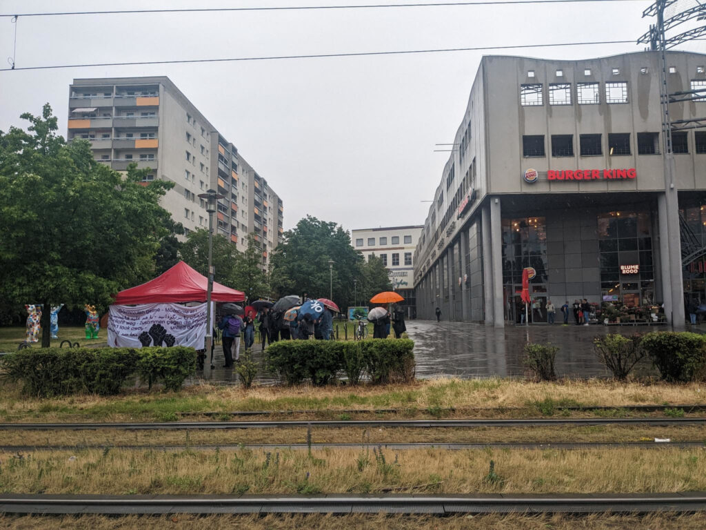 The square in front of the shopping center "Lindencenter" on a rainy day. There is a pavilion with a banner on it that says "Don't look away - together against racism" in many different languages. Next to it are some people with colorful umbrellas.